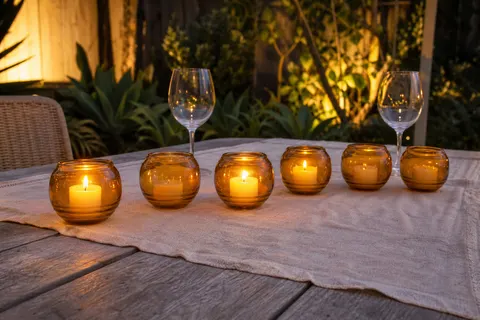 Six bougies d'extérieur en verre orange alignées sur une table de jardin en bois au crépuscule