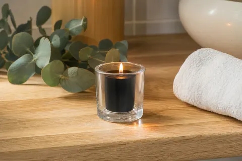 Bougie votive noire allumée dans un photophore en verre transparent, sur une étagère de salle de bain en bois clair