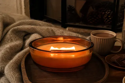Bougie à mèche en bois dans un verre elliptique ambré, flamme large allumée devant une cheminée éteinte