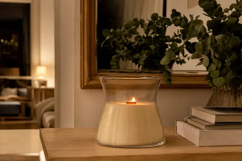 Grande bougie WoodWick en verre sablier avec mèche en bois allumée, posée sur une console en bois clair