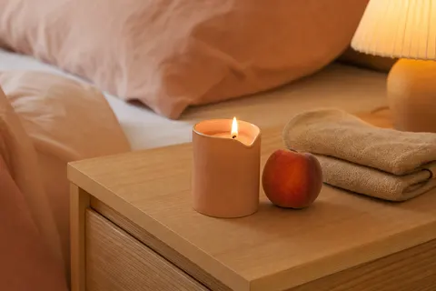 Petite bougie de massage parfum pêche à bec verseur, allumée sur une table de chevet en bois clair