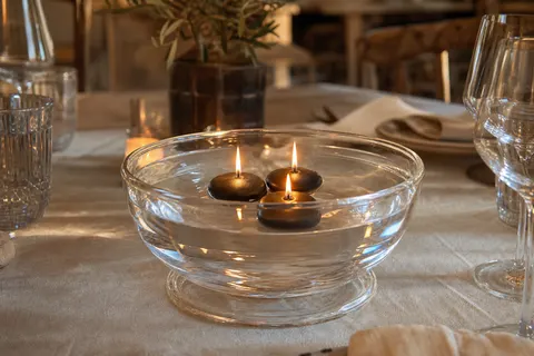 Trois bougies flottantes noires allumées dans une coupe en verre remplie d'eau, sur une table en bois dressée