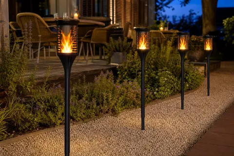 Quatre torches solaires noires à effet de flamme allumées le long d'une allée de jardin, au crépuscule