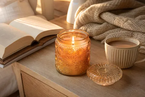Bougie en pot de verre gravé de marguerites, cire orange pâle allumée, sur une table de chevet avec un livre et du thé