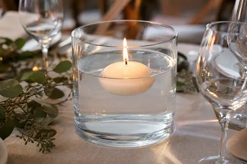 Grande bougie flottante blanche allumée dans un vase en verre rempli d'eau, sur une table nappée de lin