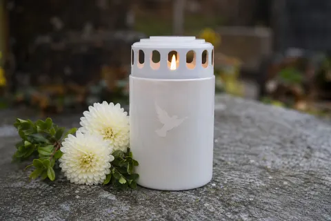 Bougie de souvenir blanche à couvercle, motif colombe, allumée sur une dalle de pierre près de fleurs blanches