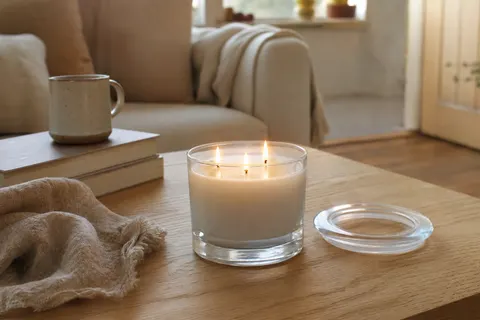 Bougie blanche à trois mèches allumée dans un pot en verre, couvercle posé sur une table basse en chêne