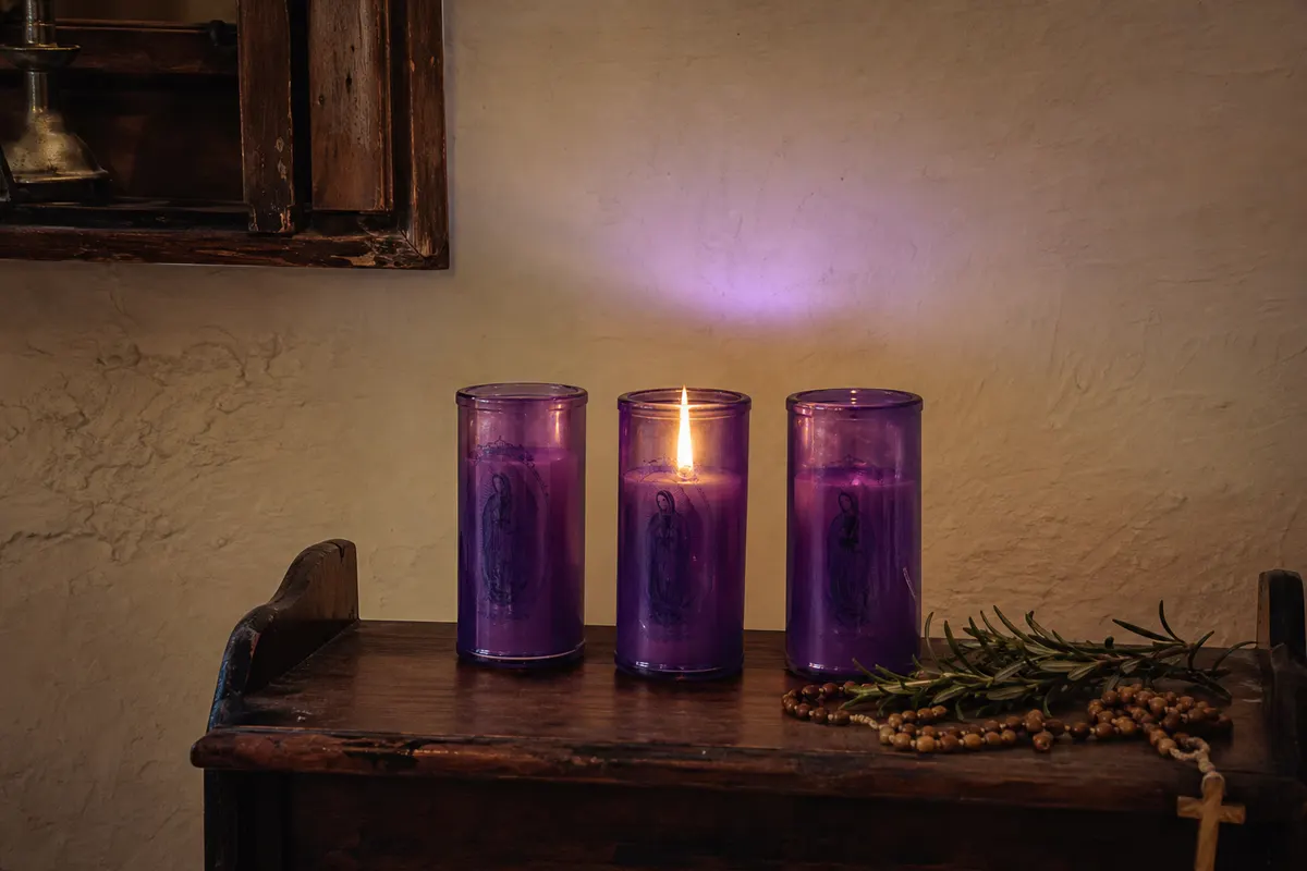 Trois bougies votives en verre violet sur une console en bois, une flamme allumée près d'un chapelet