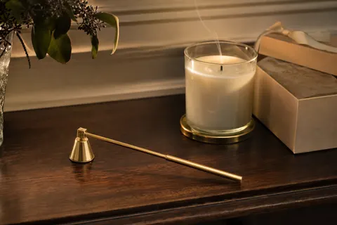 Éteignoir doré à long manche et cloche articulée posé sur une commode près d'une bougie qui vient d'être éteinte