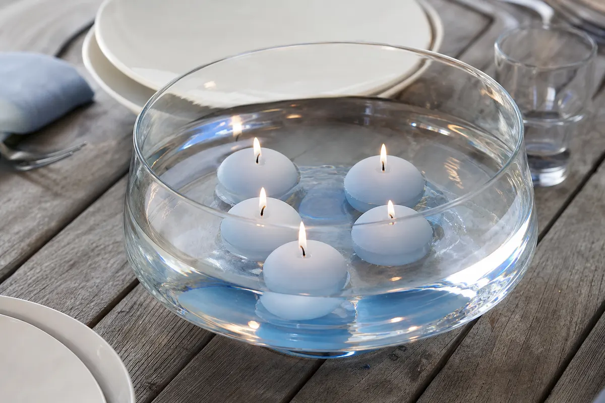 Cinq bougies flottantes bleu poudré allumées dans une coupe en verre, sur une table de jardin en bois