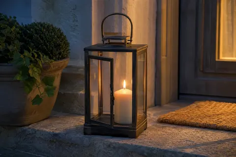 Lanterne carrée en métal noir et cuivre, vitrée, avec une bougie pilier allumée, sur une table de jardin en bois