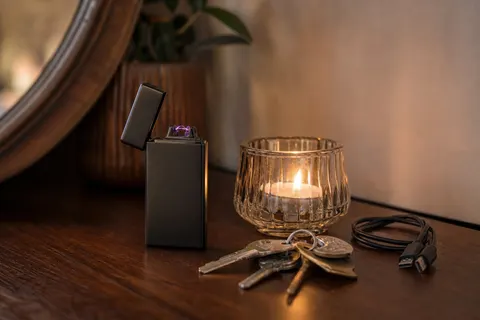 Briquet électrique noir givré à double arc posé sur une commode en bois près d’une bougie chauffe-plat allumée