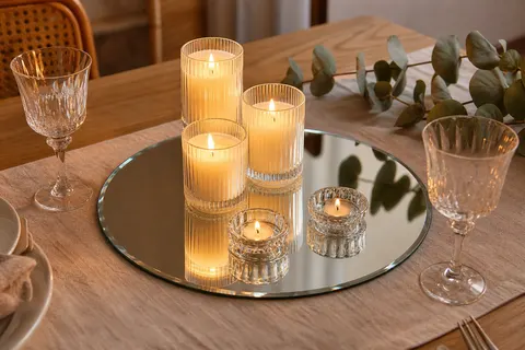 Plateau rond en verre miroir avec bougies en pot allumées reflétées, sur une table en bois clair