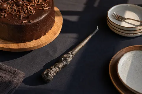 Bougie sculptée en forme de baguette de sorcier posée sur une nappe bleu nuit à côté d'un gâteau au chocolat