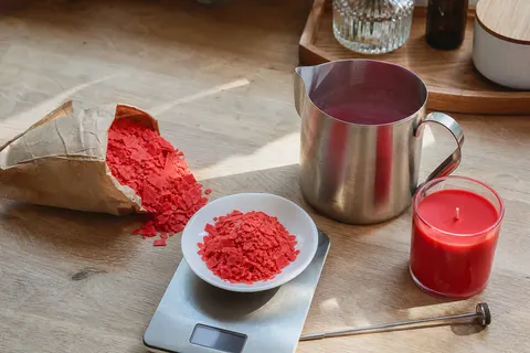 Flocons de colorant rouge pour bougies dans une coupelle sur une balance, avec pichet de coulée en inox