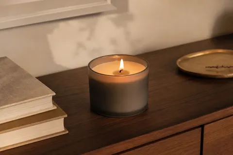 Bougie en pot taupe mat à mèche allumée, posée sur une commode en noyer près de livres reliés et d'un plateau en laiton