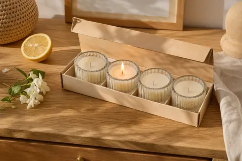 Coffret ouvert de quatre bougies en cire de soja dans des pots en verre, dont une allumée, sur une commode