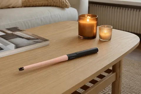 Allume-bougie électrique à arc rose pâle posé sur une table basse en bois près de deux bougies en pot de verre