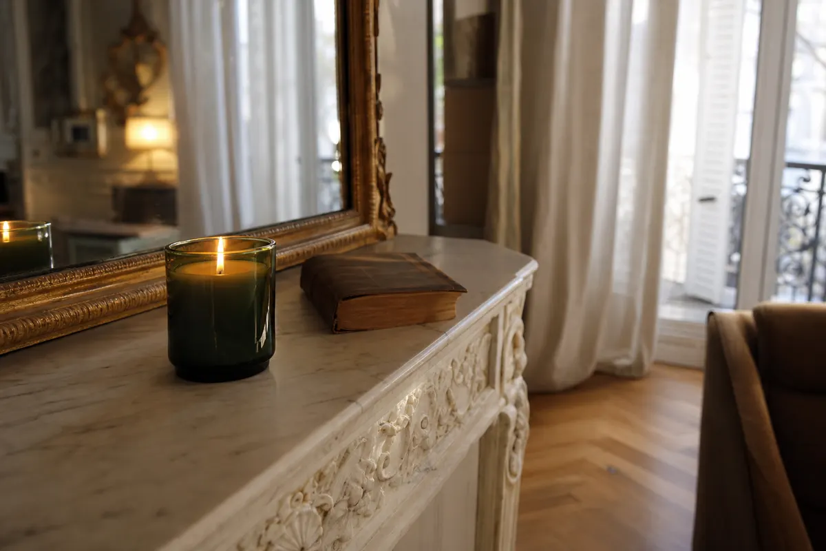 Bougie parfumée allumée dans un verre soufflé vert, posée sur une cheminée en marbre d'un salon parisien